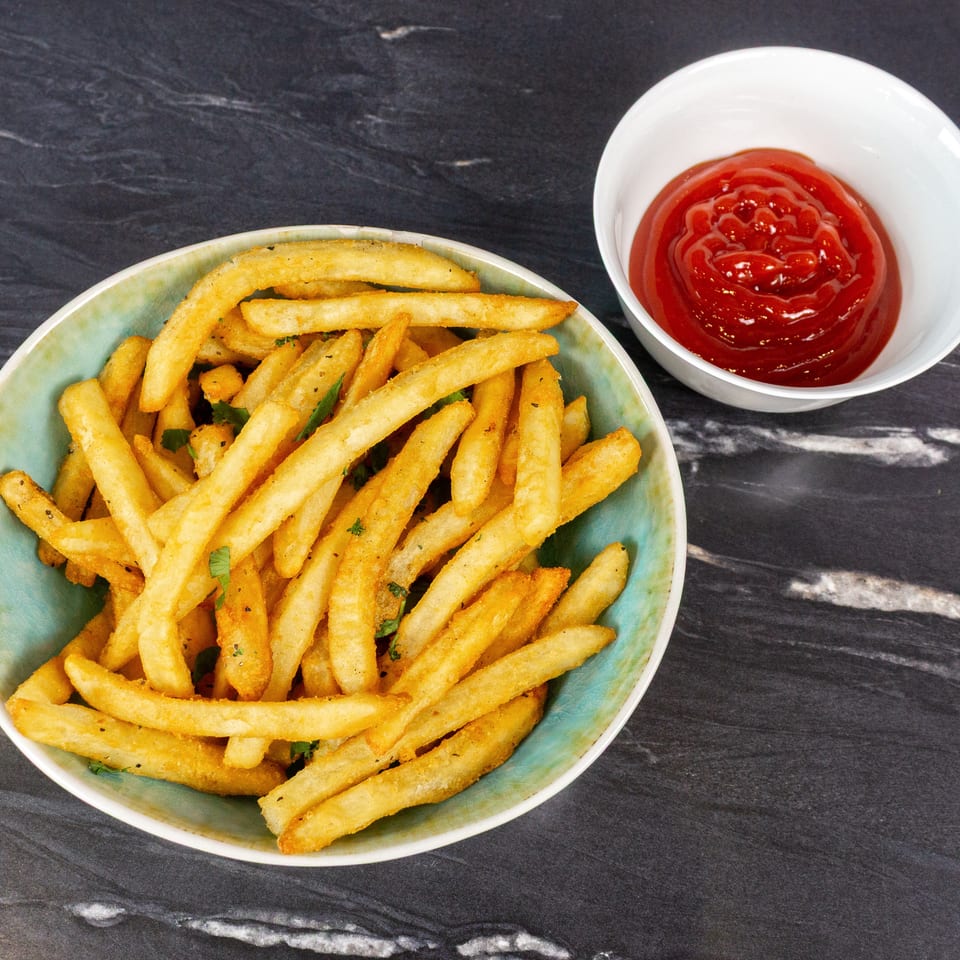 Bowl of golden seasoned fries with fresh herbs beside a small dish of ketchup on a dark marble tabletop.