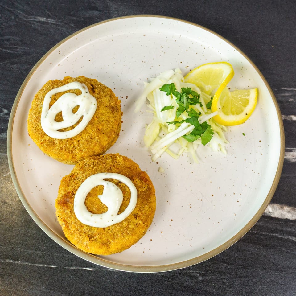 Two golden breaded patties topped with creamy sauce, served with shredded cabbage, parsley, and lemon wedges on a plate.