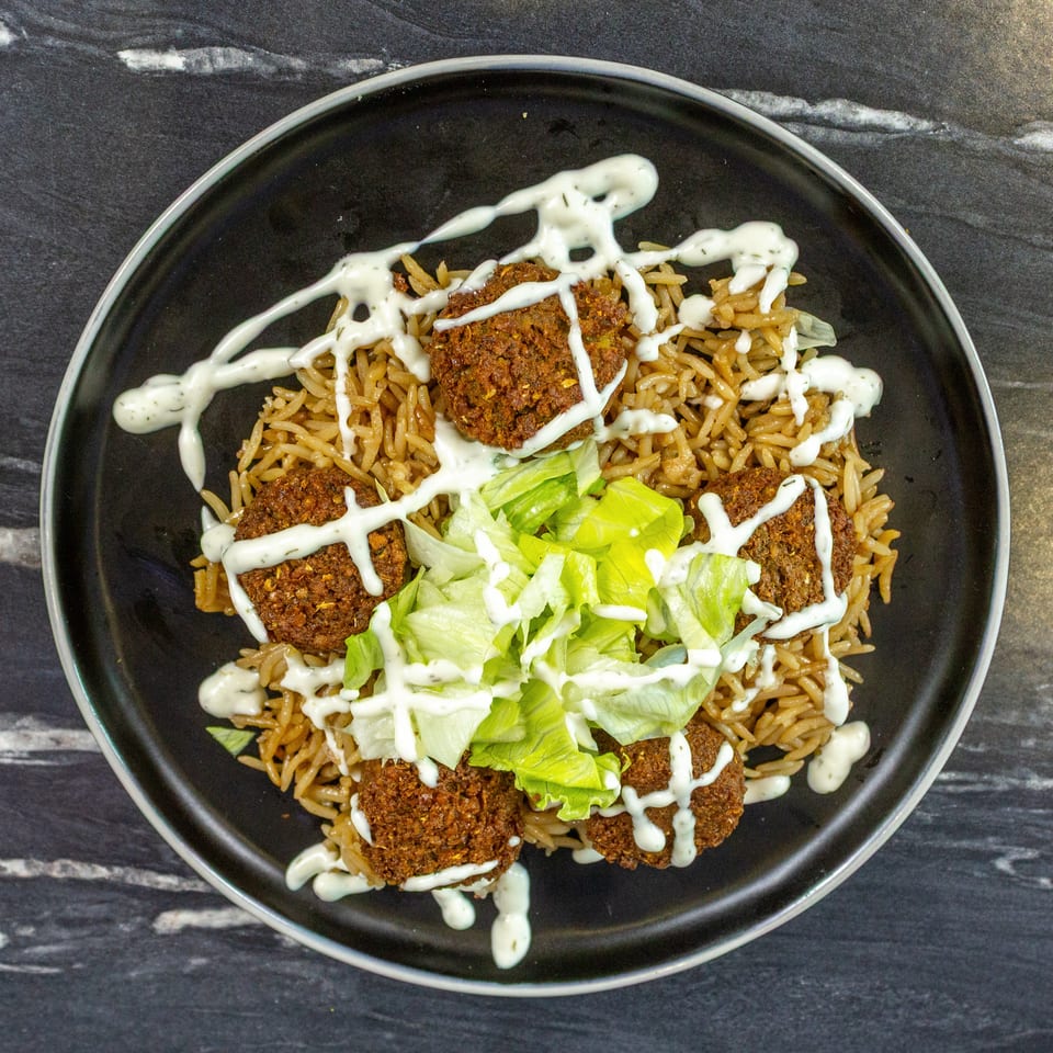Black plate of spiced rice topped with falafel, lettuce, and creamy white sauce.
