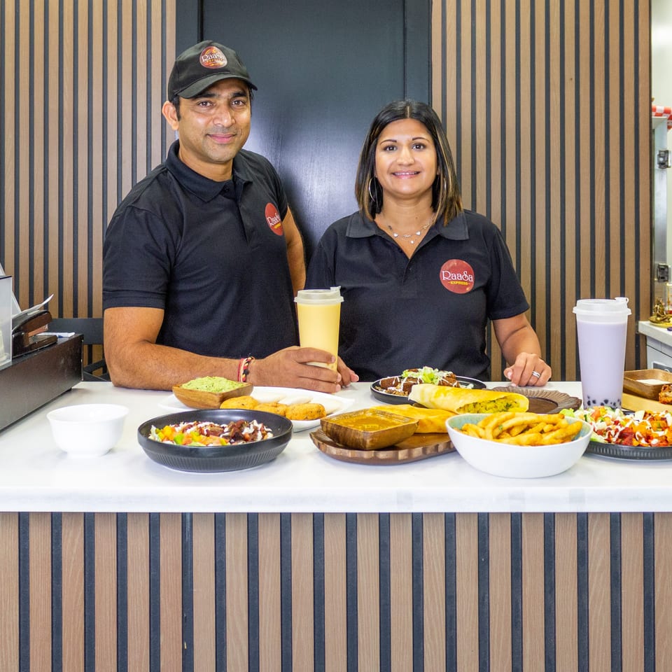 Two Raada Express staff members pose behind a counter displaying colorful Indian dishes, wraps, fries, desserts, and drinks.