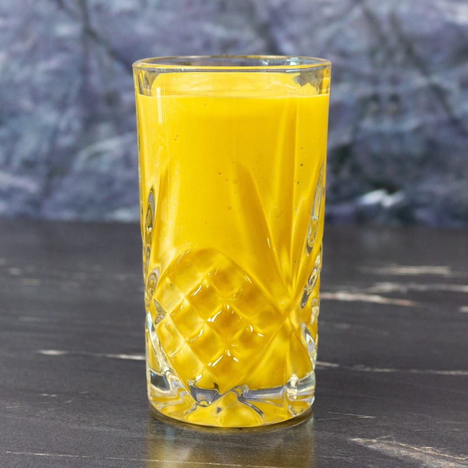 Glass of bright yellow juice on a dark tabletop, with a softly blurred gray background.