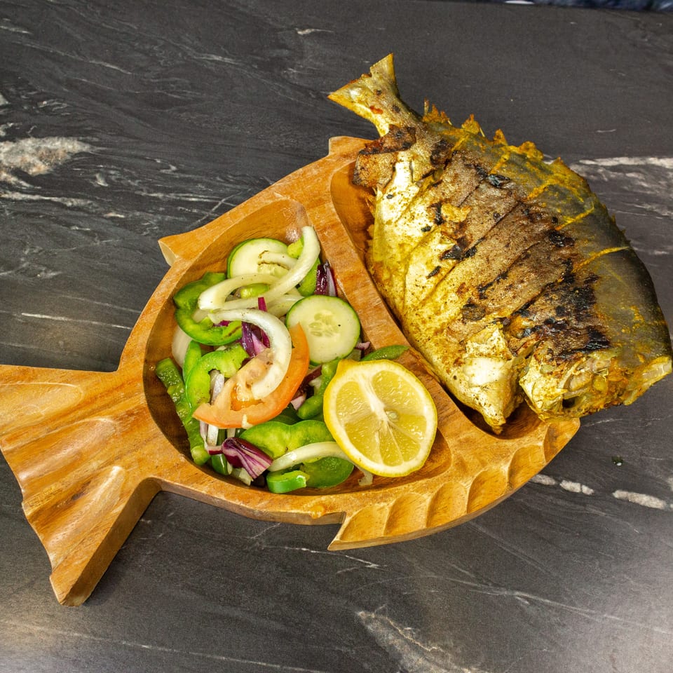 Grilled whole fish served on a wooden platter with sliced lemon and a fresh salad of cucumbers, peppers, onions, and tomatoes.