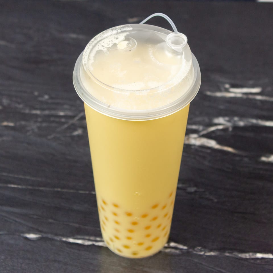 Yellow bubble tea with tapioca pearls and foamy topping in a clear takeaway cup on a dark marble surface.