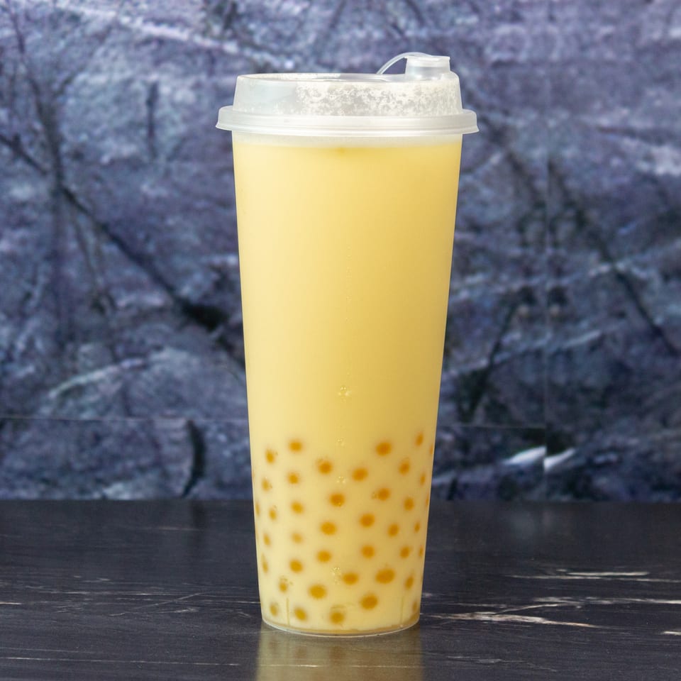 Yellow bubble tea with tapioca pearls in a clear lidded cup on a dark table.