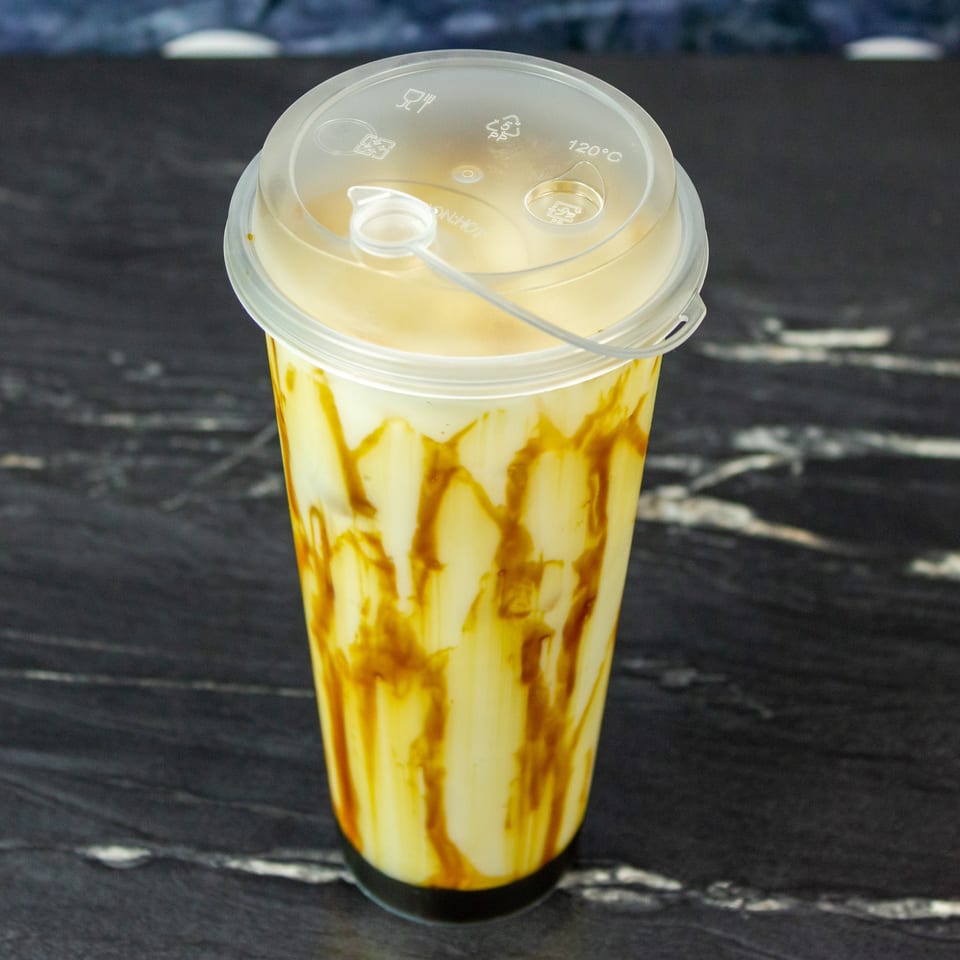 Caramel-drizzled creamy yellow beverage in a clear plastic cup with a lid and straw, on a dark wooden table.