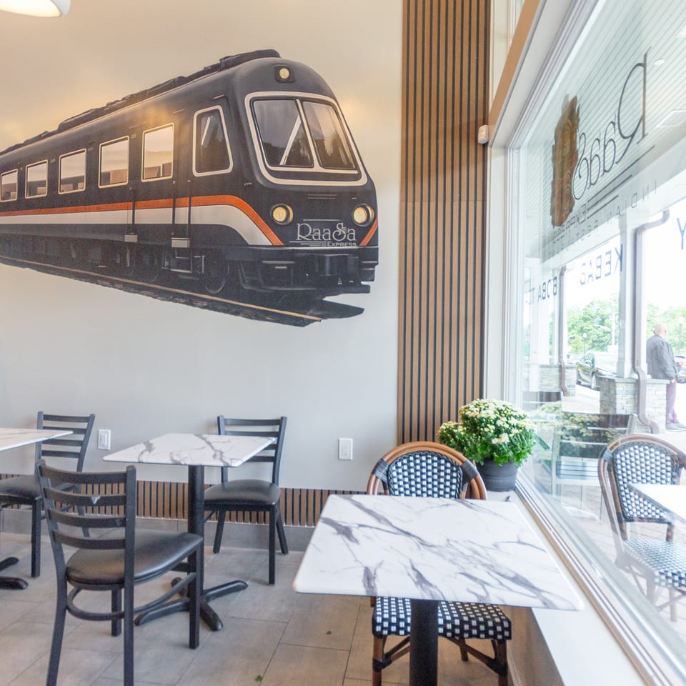 Restaurant dining area with marble tables, woven chairs, large windows, and a decorative Raaca Express train mural.