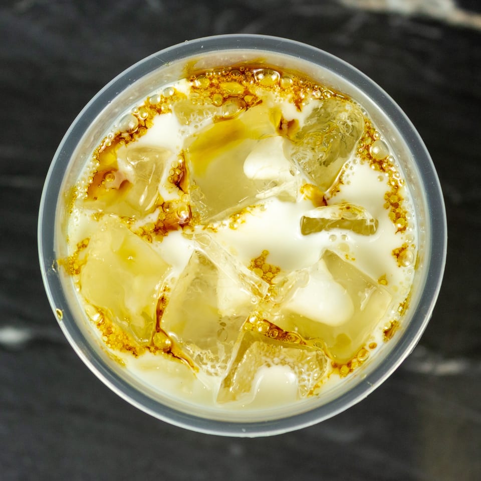 Top-down view of a creamy golden iced beverage with ice cubes and bubbles in a clear plastic cup.