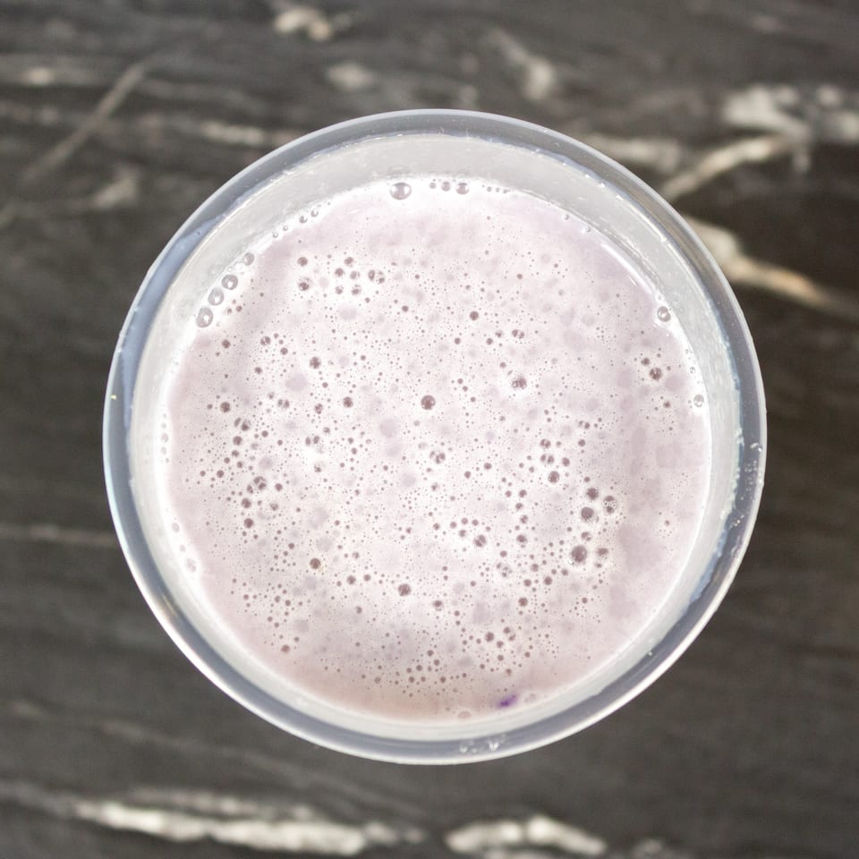 Top-down view of a clear glass filled with pale purple, frothy beverage on a dark wooden table.