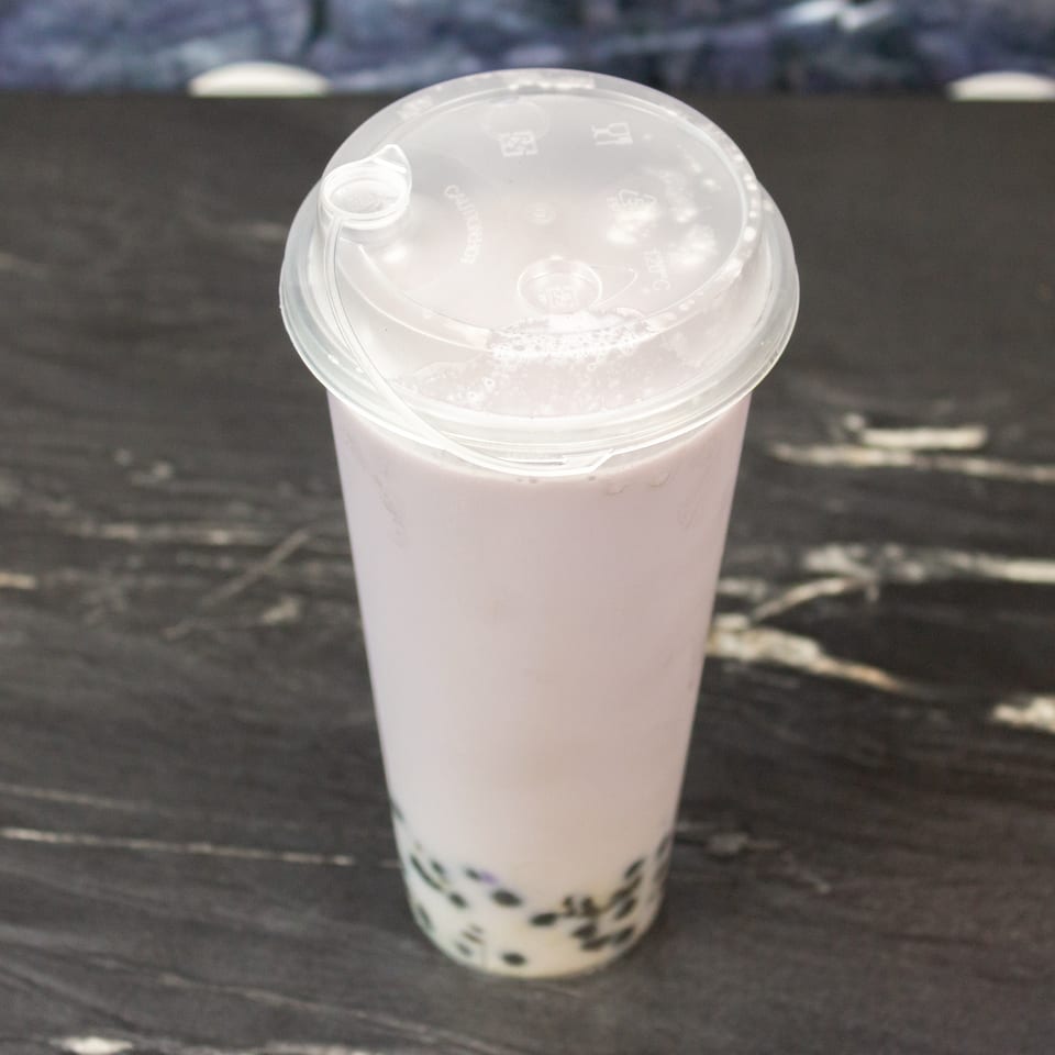 Pale milk tea with dark tapioca pearls in a clear takeaway cup on a dark wooden table.