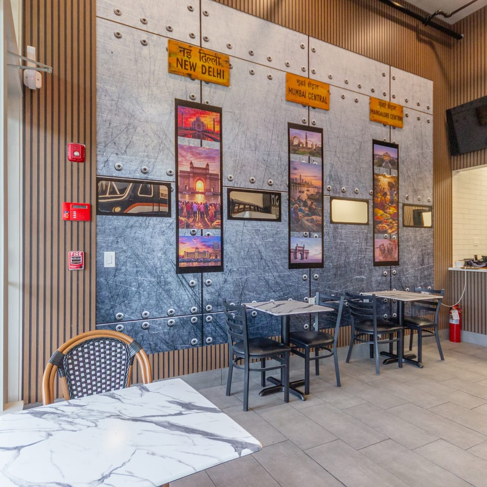 Restaurant dining area with marble tables, dark chairs, striped walls, and travel-themed artwork labeled New Delhi, Mumbai Central, and Bangalore Central.