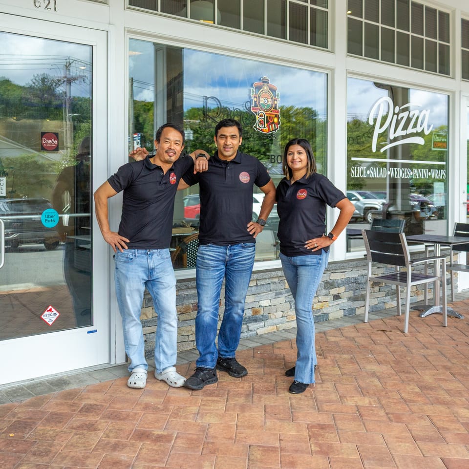 Three Raasa Pizza restaurant staff members pose outside the storefront beside patio seating and windows advertising pizza, salads, wedges, panini, and wraps.